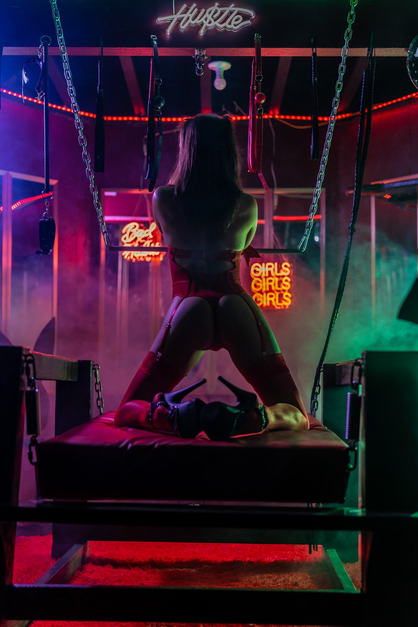Woman in red lingerie kneeling in a neon-lit boudoir room with moody red and blue lighting.