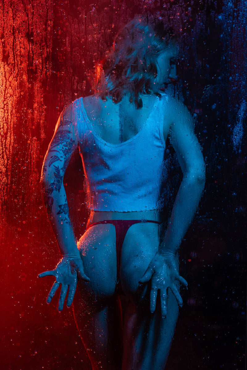 Artistic boudoir silhouette behind a steamy shower glass panel with dramatic red and blue backlighting, creating a moody and modern cinematic look in Wisconsin.