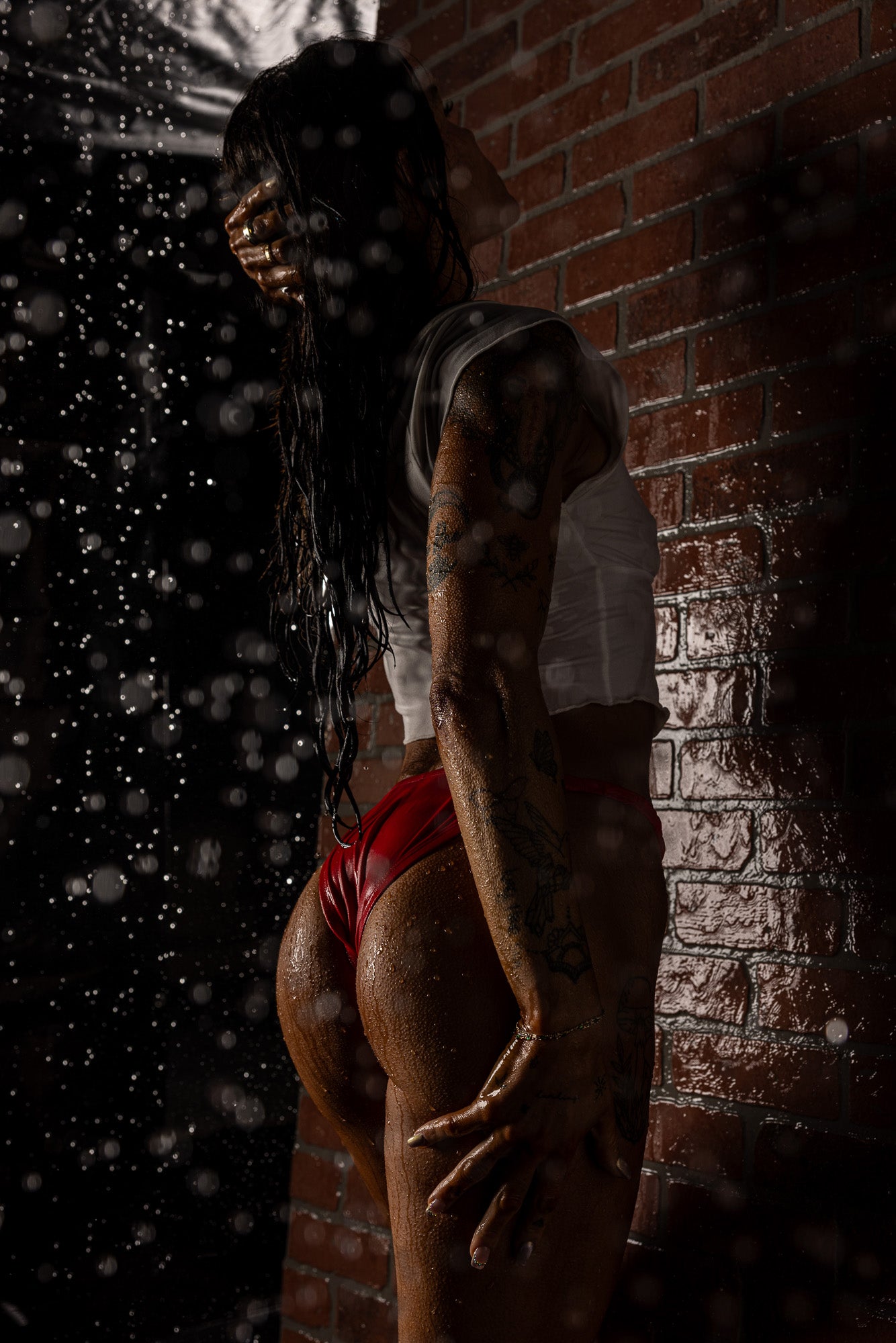 Wet-look boudoir portrait featuring a woman in red lingerie posing against a brick wall, captured with dramatic lighting to emphasize shine, texture, and mood.