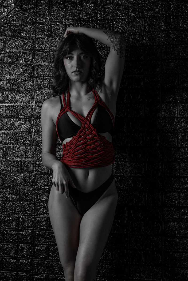 Artistic boudoir portrait featuring red shibari rope against a matte black wall, with the model standing in a strong vertical pose under dramatic low-key studio lighting.